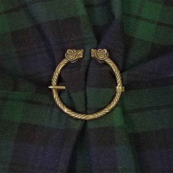 A Boar Penannular Brooch with intricate bronze Celtic designs on its terminals is laid on a dark green and blue plaid fabric.