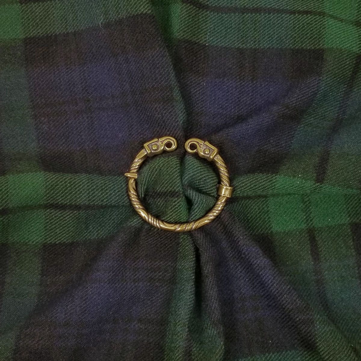 A Falcon Penannular Brooch fastens a piece of green and blue tartan fabric, reminiscent of ancient falcon motifs often seen in historical designs.