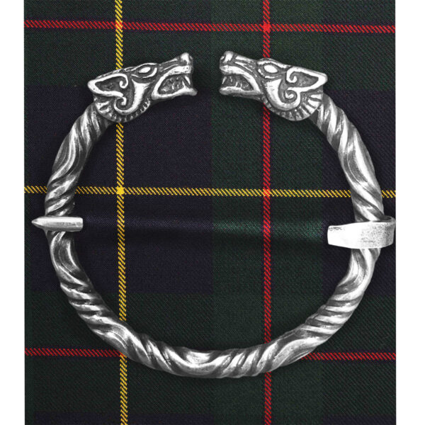A Wolf Penannular Brooch in silver, featuring a Celtic design with two wolf heads on each end, is placed on plaid fabric, evoking the look of a traditional brooch.