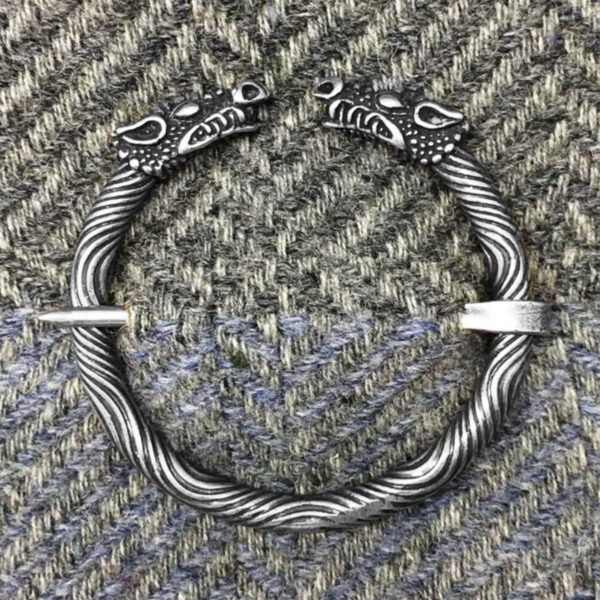 The Celtic Dragon Penannular Brooch, featuring two intricately designed dragon heads on each end, is displayed on a herringbone-patterned fabric background.