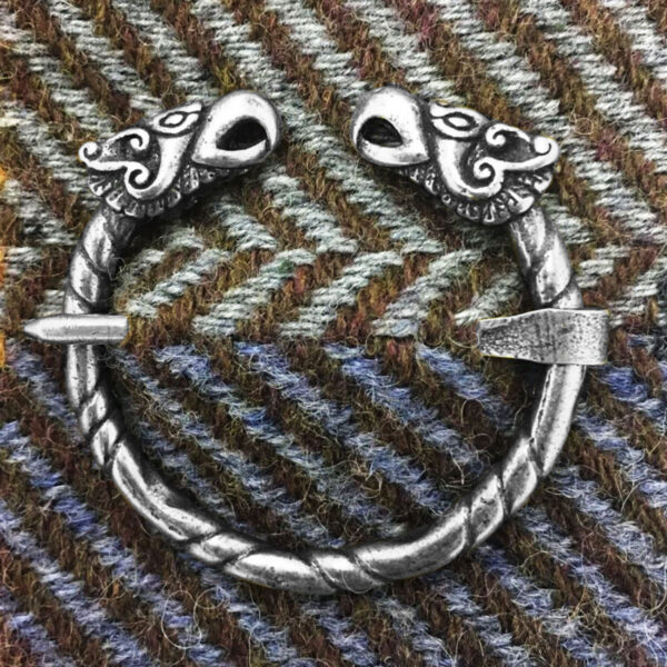The Griffin Penannular Brooch, featuring a circular silver design adorned with two dragon heads and griffin-inspired artistry, is placed on a background of herringbone patterned fabric.