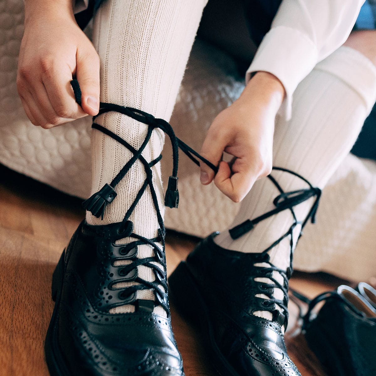 A person ties the Premium Ghillie Laces of black dress shoes while wearing white knee-high socks. The setting appears to be indoors on a wooden floor, adding a touch of premium elegance to the scene.
