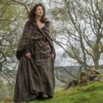 A person with curly hair stands outdoors wearing a long, brown Welsh Tartan Earasaid - 13oz Medium Weight Premium Wool. They are surrounded by trees and greenery.
