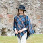 A woman in a Light Weight 11oz Premium Wool Tartan Poncho, standing in front of a stone wall.