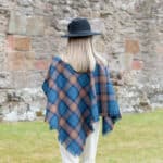 A woman wearing a Light Weight 11oz Premium Wool Tartan Poncho made from light weight 11oz premium wool fabric in front of a stone wall.