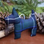 A Tartan Scottie Dog Ornament sits on a wooden surface, surrounded by pine cones and greenery in the background.