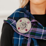 A close-up of a person wearing a blue and purple tartan scarf fastened with one of our Crest and Gem Plaid Brooches, adorned with four red gems and featuring intricate patterns around a central thistle motif.