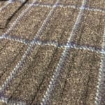 Close-up of a Twill Tweed Kilt showcasing a grey wool textile with a blue and white plaid pattern. The fabric appears textured and warm, reminiscent of traditional tweed.