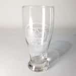 The MacKenzie Clan Crest 19 oz Pilsner Glass, made from clear glass and etched with the design "Na Muckarach," stands elegantly on a white surface casting a faint shadow.