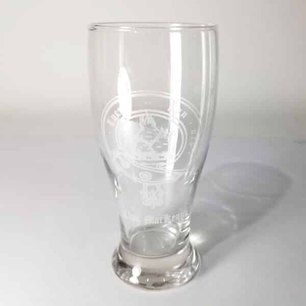 The MacKenzie Clan Crest 19 oz Pilsner Glass, made from clear glass and etched with the design "Na Muckarach," stands elegantly on a white surface casting a faint shadow.