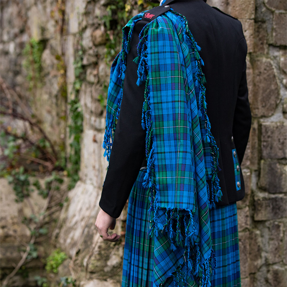 A man in a Welsh Tartan Medium Weight Premium Wool Fly Plaid kilt standing next to a stone wall.