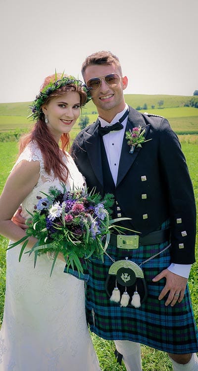 A bride and groom in a kilt standing in a field.