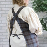 A woman wearing a Tartan Cross Back Apron - Homespun Wool Blend in front of a brick wall.