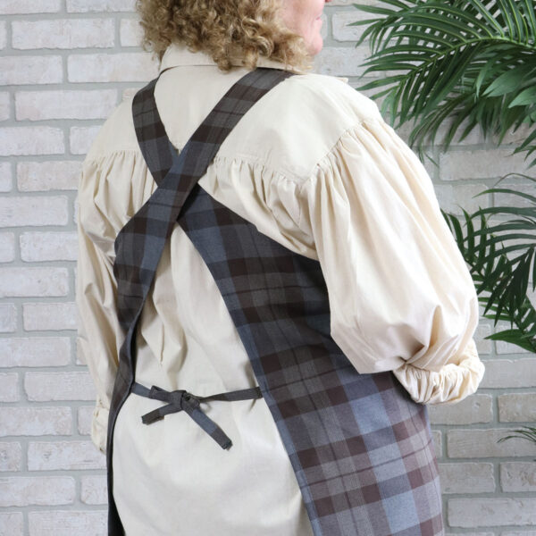A woman wearing a Tartan Cross Back Apron - Homespun Wool Blend in front of a brick wall.