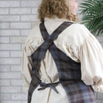 A woman wearing a Tartan Cross Back Apron - Homespun Wool Blend standing in front of a brick wall.