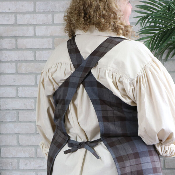 A woman wearing a Tartan Cross Back Apron - Homespun Wool Blend standing in front of a brick wall.