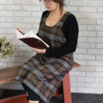 A woman wearing a Tartan Cross Back Apron - Homespun Wool Blend sits on a bench, engrossed in her book.
