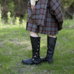 A woman in a plaid kilt standing in the grass, wearing Black Premium Leather Knee-High Boots.