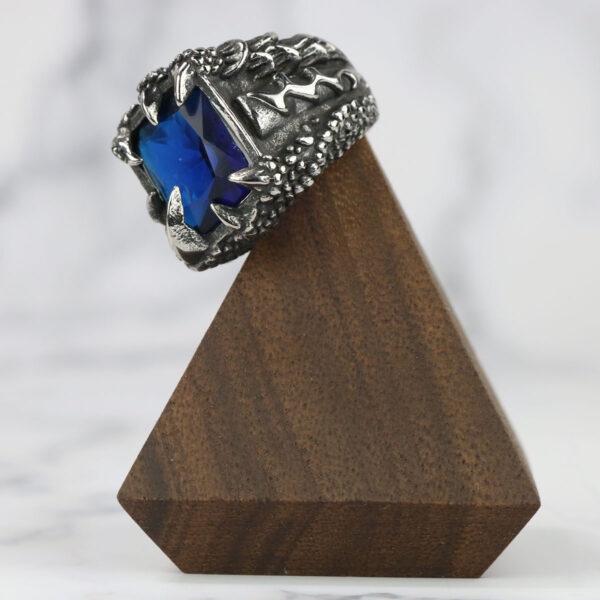 Description: A Dragon Claw Stainless Steel Ring with a blue stone on top of a wooden base.