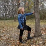 A young girl wearing a Wilderness Kilts for Kids, standing next to a tree.
