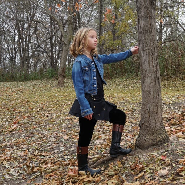 A young girl wearing a Wilderness Kilts for Kids, standing next to a tree.