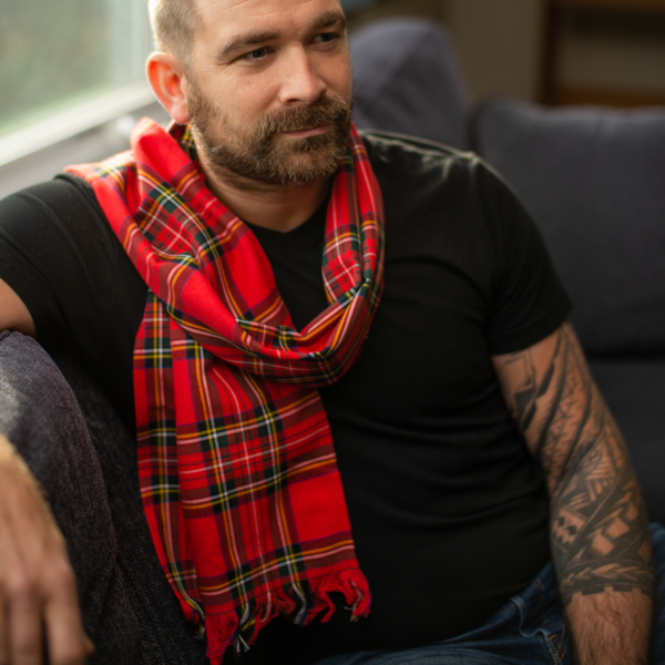 A man sitting on a couch wearing a red Tartan Scarf - 13oz Premium Wool, accessorized with a medium weight scarf.