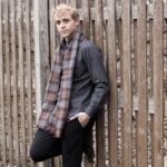 A young man leaning against a wooden fence wearing a plaid scarf and an OUTLANDER Golf Cap Authentic Premium Wool Tartan.