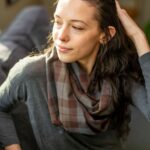 A woman is sitting on a couch wearing a Tartan Infinity Scarf - OUTLANDER Wool Free.