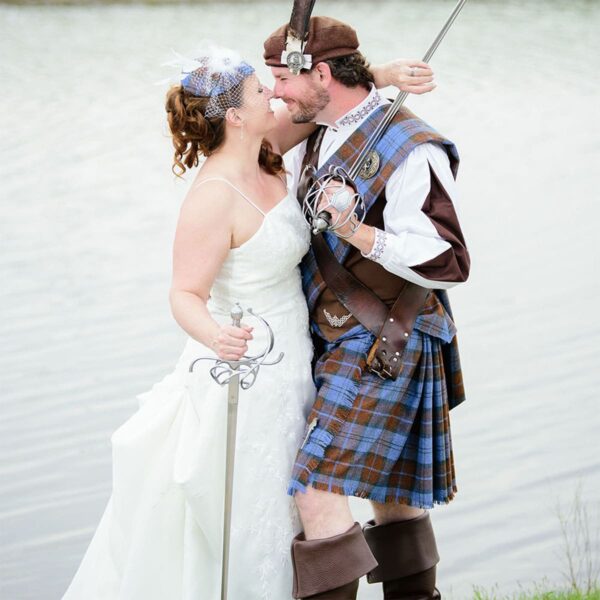 A bride and groom dressed in kilts are standing next to a body of water.