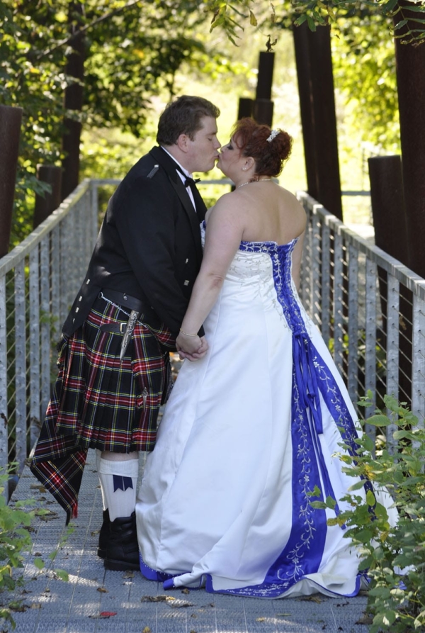 A couple in kilts, dressed in the Prince Charlie Formal PLATINUM Package, share a romantic kiss on a bridge.