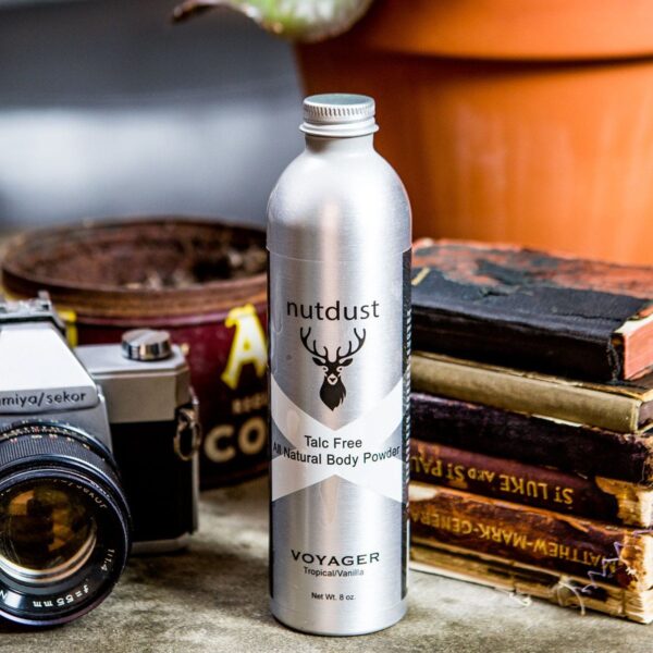 A bottle of Nutdust - Voyager body wash sits on a table next to a camera and books.