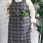 A woman wearing a Tartan Cross Back Apron - Homespun Wool Blend with a plant in it.