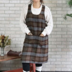 A woman wearing a Tartan Cross Back Apron - Homespun Wool Blend standing in front of a brick wall.