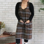 A woman wearing a Tartan Cross Back Apron - Homespun Wool Blend standing next to a brick wall.