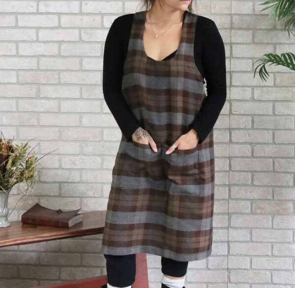 A woman wearing a Tartan Cross Back Apron - Homespun Wool Blend standing next to a brick wall.