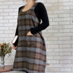 A woman wearing a Tartan Cross Back Apron - Homespun Wool Blend standing in front of a brick wall.