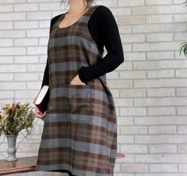 A woman wearing a Tartan Cross Back Apron - Homespun Wool Blend standing in front of a brick wall.