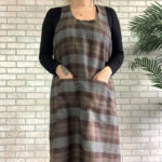 A woman wearing a Tartan Cross Back Apron - Homespun Wool Blend standing in front of a brick wall.