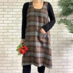 A woman wearing a Tartan Cross Back Apron - Homespun Wool Blend standing next to a brick wall.