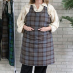 A woman wearing a Tartan Cross Back Apron - Homespun Wool Blend in front of a brick wall.