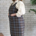 A woman wearing a Tartan Cross Back Apron - Homespun Wool Blend in front of a brick wall.