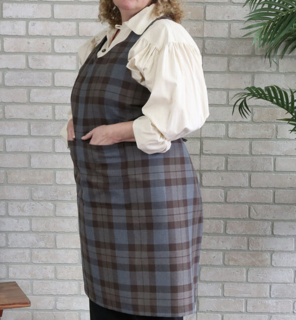 A woman wearing a Tartan Cross Back Apron - Homespun Wool Blend in front of a brick wall.