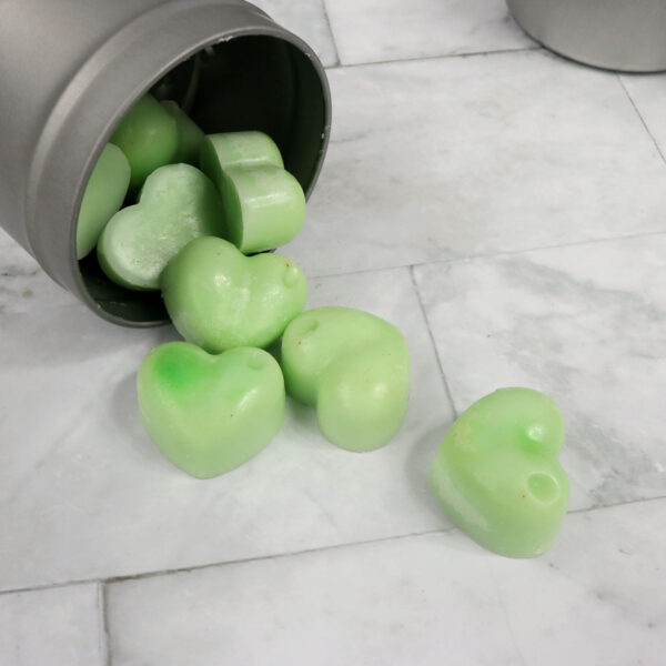 Homemade green heart soap in a metal container on a marble floor. → Homemade candles in a metal container on a marble floor.