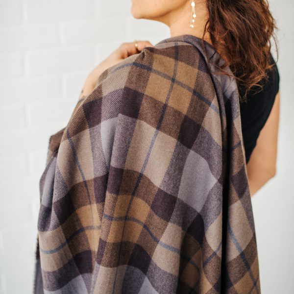 A woman wearing an OUTLANDER Wrap Premium Lambswool Tartan in a brown and tan plaid blanket.