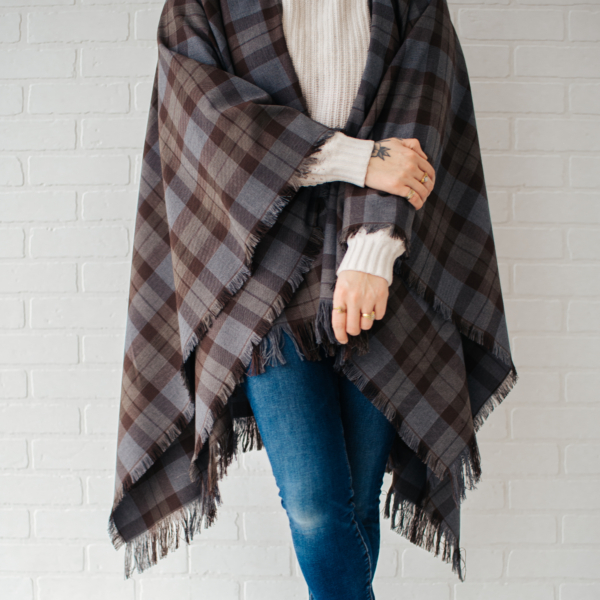 A woman wearing an OUTLANDER Wrap Premium Lambswool Tartan standing in front of a brick wall.